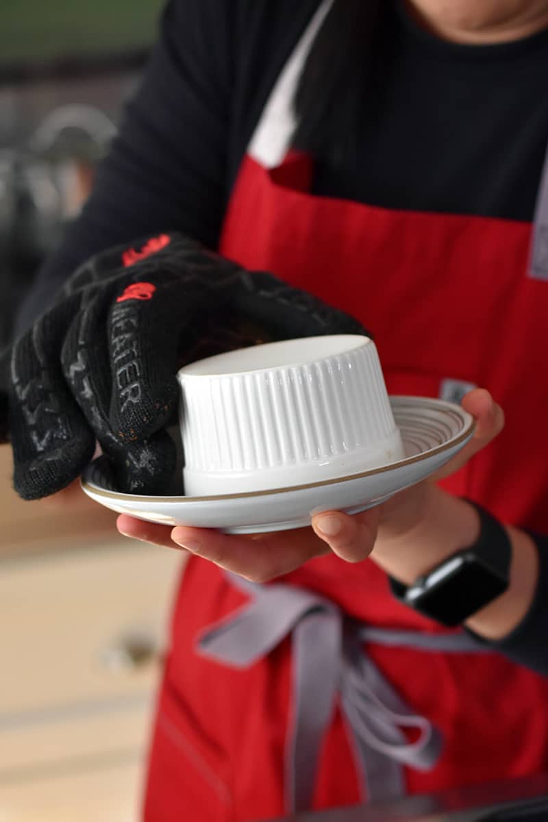 A person in a red apron is removing a ramekin off a plate with a kevlar glove.