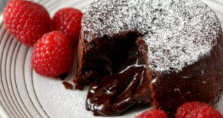 A closeup shot of a white plate with a chocolate lava cake opened to show the gooey center. There is powdered keto confectioners sugar on top and raspberries on the side.