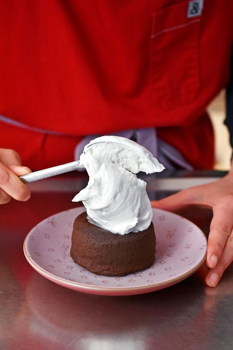 A person in a red apron is putting a large dollop of whipped coconut cream on top of a gluten-free paleo chocolate lava cake
