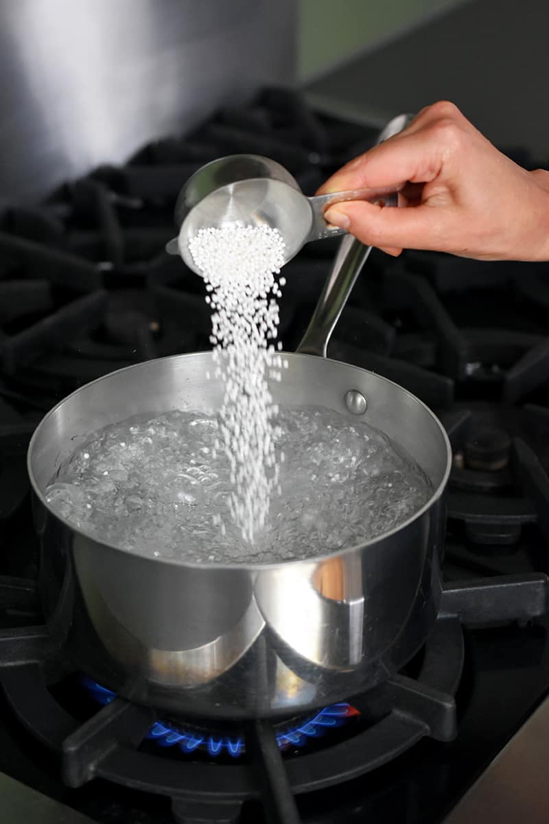 A hand is adding small tapioca pearls into a saucepan filled with boiling water.