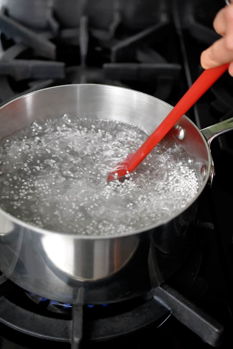 A closeup of a large saucepan filled with boiling water and small tapioca pearls. A red spatula is stirring the contents of the pan.