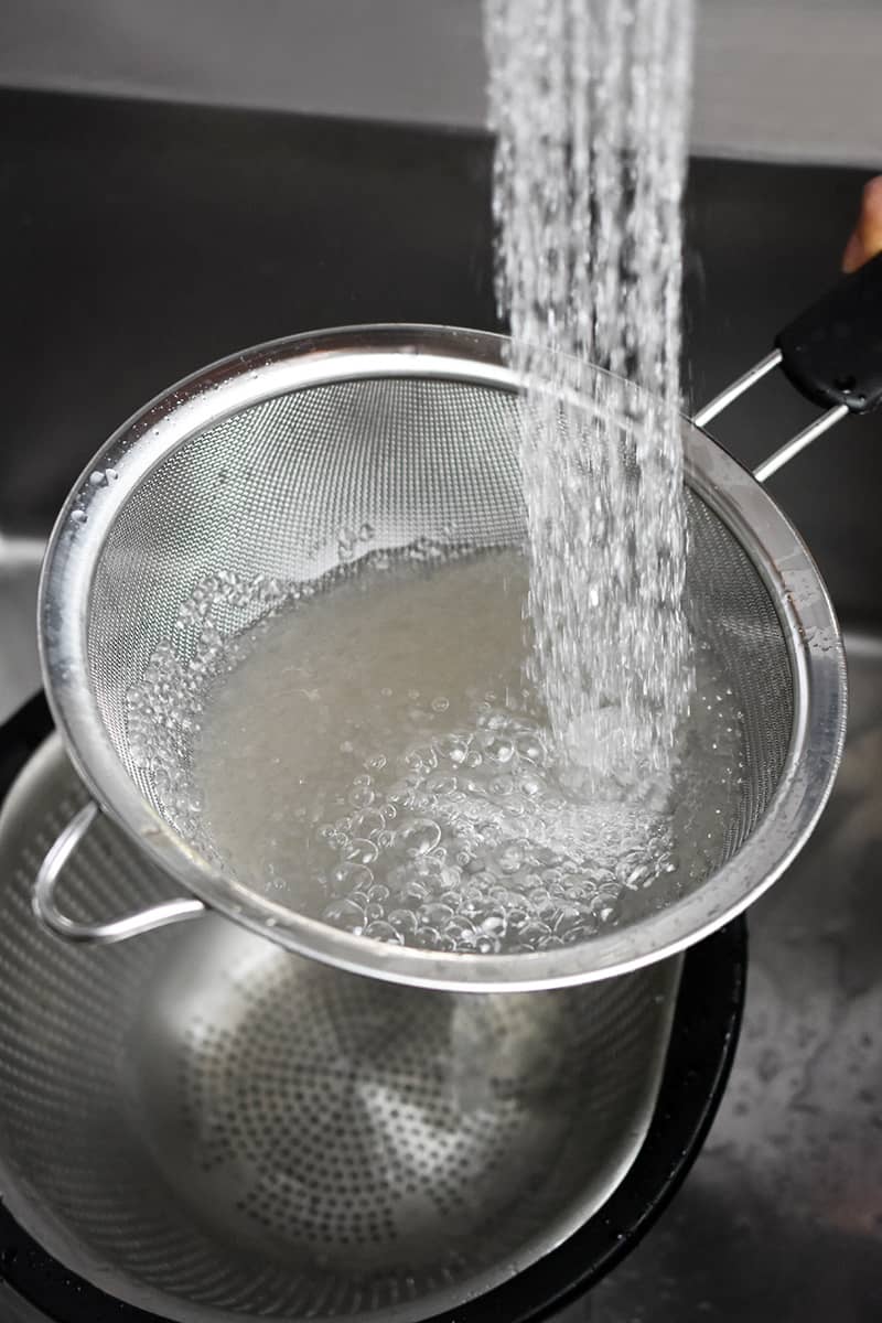 Running water is being poured into a fine mesh sieve filled with cooked small tapioca pearls.