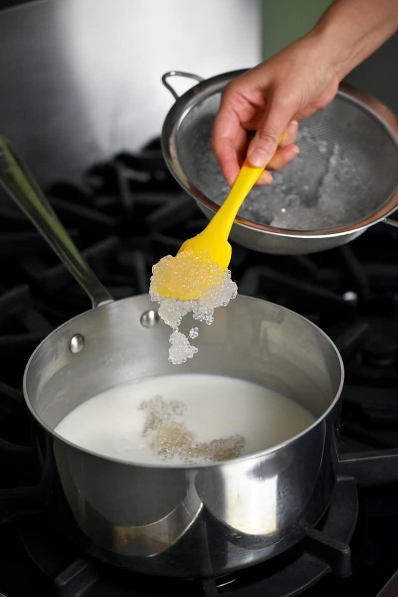 A yellow spatula is spooning cooked small tapioca pearls into a saucepan filled with sweetened coconut milk.