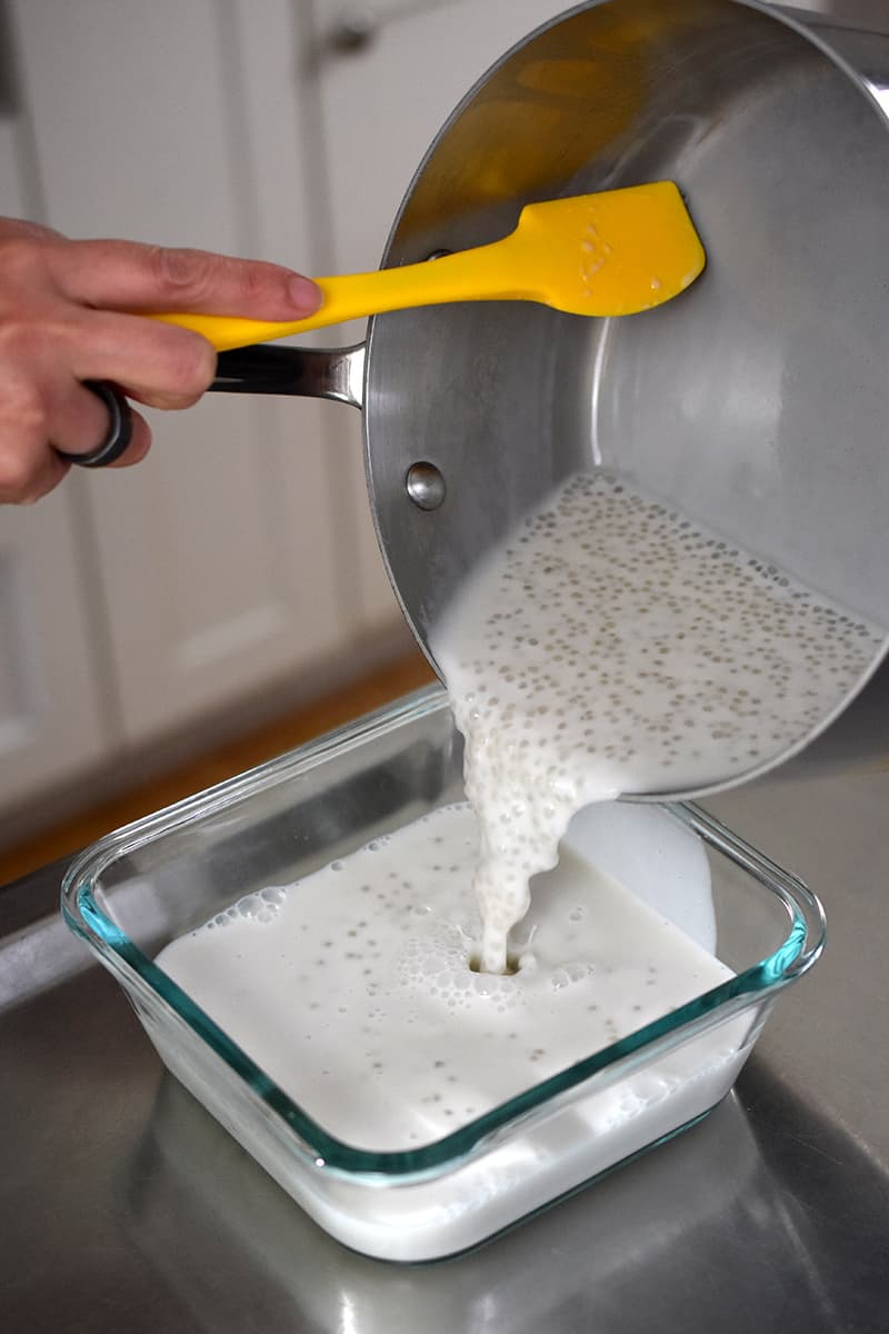 A shot of someone transferring coconut milk tapioca pudding from a saucepan into a glass square container.
