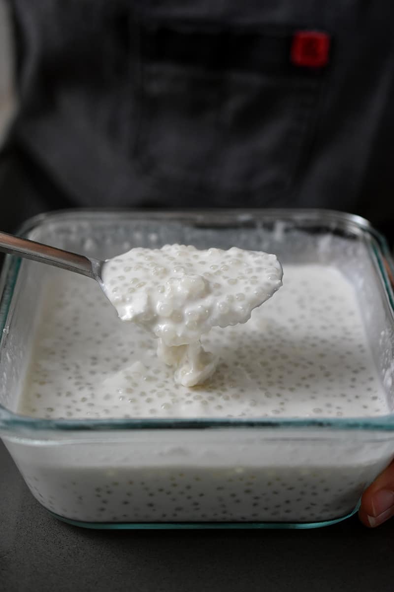 A spoon is lifting up a spoonful of coconut milk tapioca from a square glass container.