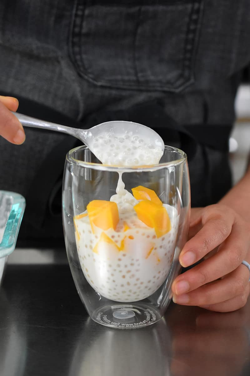 A front shot of someone layering coconut sago with diced mango in a clear glass cup.