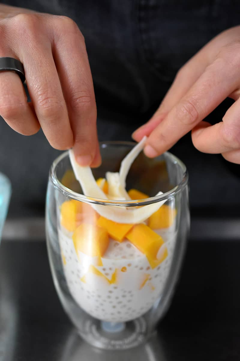 Two hands adding toasted coconut on top of clear glass filled with mango sago, a coconut tapioca pudding with diced mango