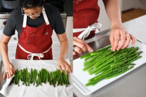 Someone in a red apron is drying asparagus with a white kitchen towel and then arranging them on a rectangular white serving platter.