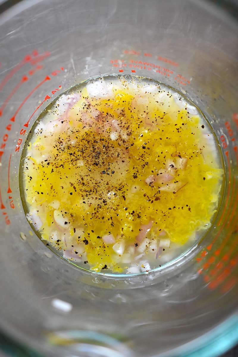 An overhead shot closeup shot of a liquid measuring cup filled with minced shallots, lemon zest, lemon juice, champagne vinegar, salt, and pepper