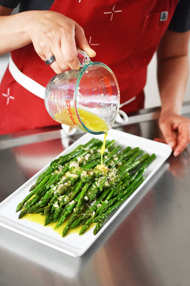 Someone in a red apron is pouring a lemon and shallot vinaigrette on top of cooked asparagus on a white rectangular plate.