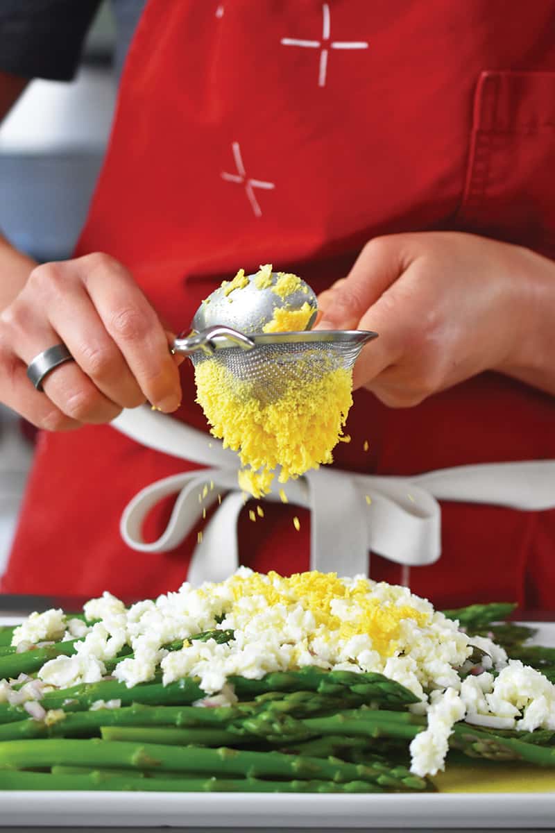 Someone in a red apron is pushing cooked egg yolks though a small mesh sieve onto a plate of asparagus topped with egg whites.