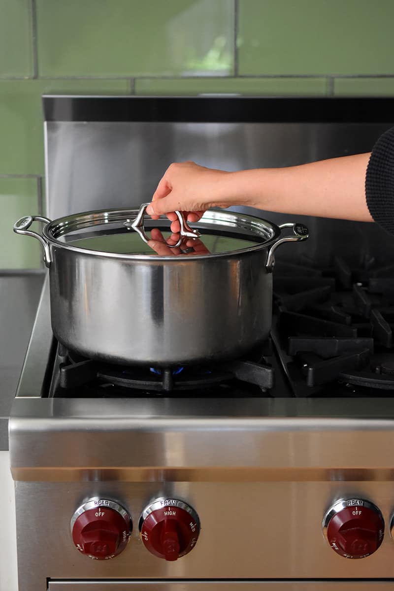 A hand is putting a lid on a large stockpot filled with water on a stovetop.