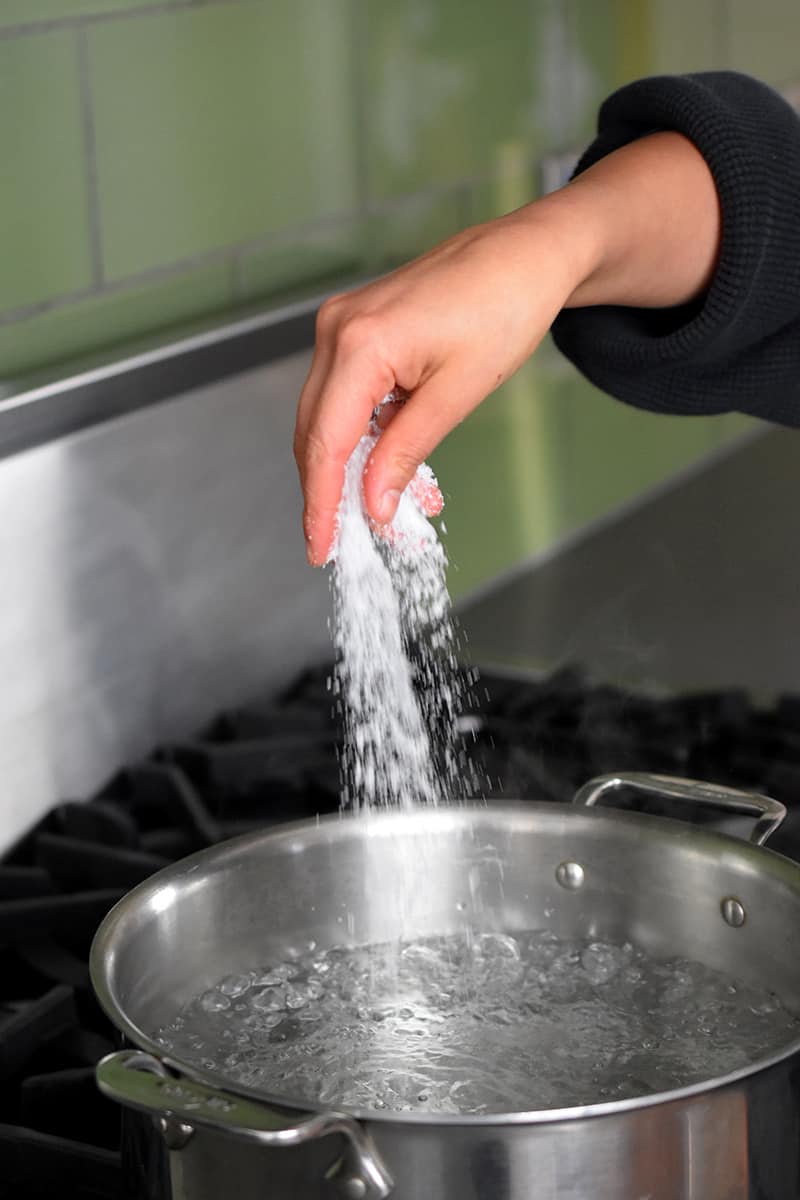 A hand is adding a large pinch of salt into a pot of boiling water on a stovetop.