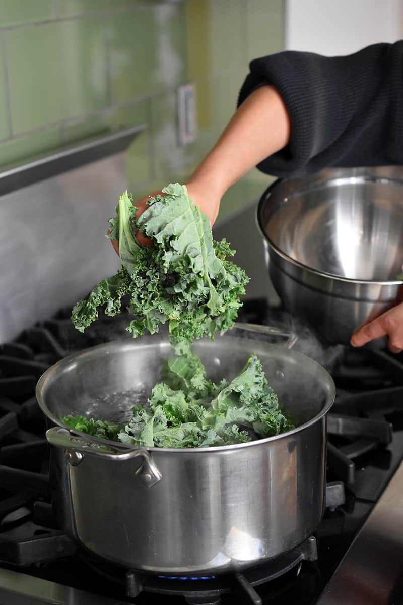 A hand is adding curly kale into a open pot of salted water on stovetop.