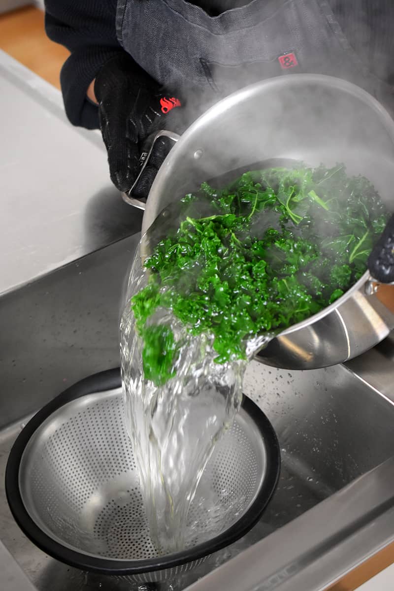 Pouring blanched kale from a stockpot into a colander in a sink.