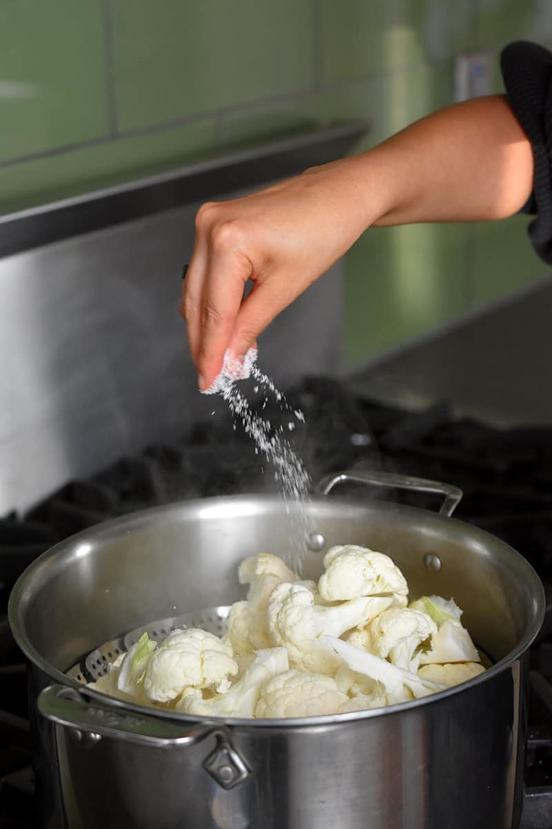 A hand is sprinkling salt into an open stockpot filled with cauliflower in a steamer.