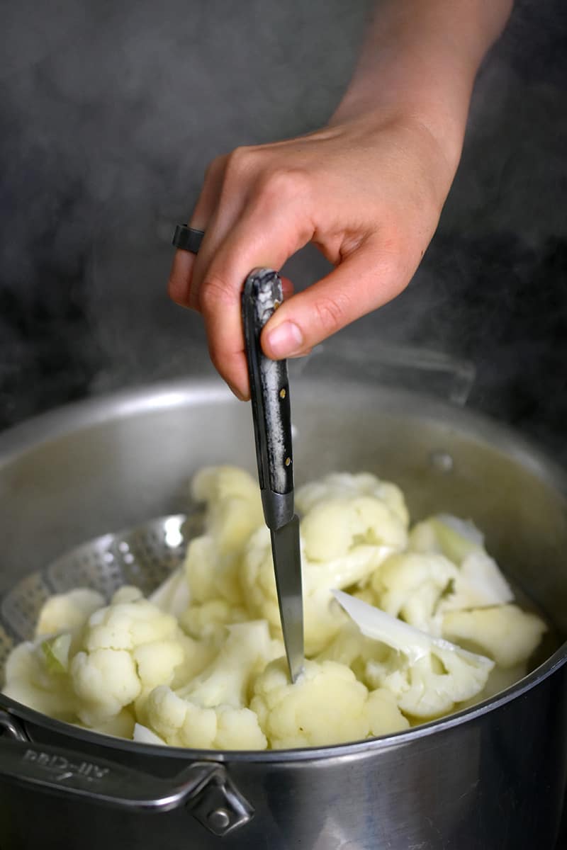 A hand is using a paring knife to pierce a pice of steamed cauliflower in an open stockpot to see if it is ready.