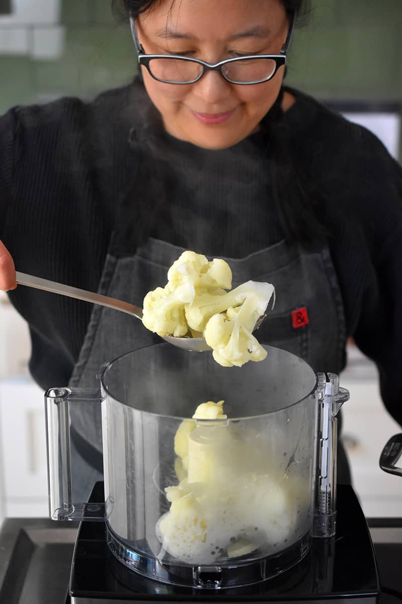 A person in a gray apron is adding cooked cauliflower florets into an open food processor.