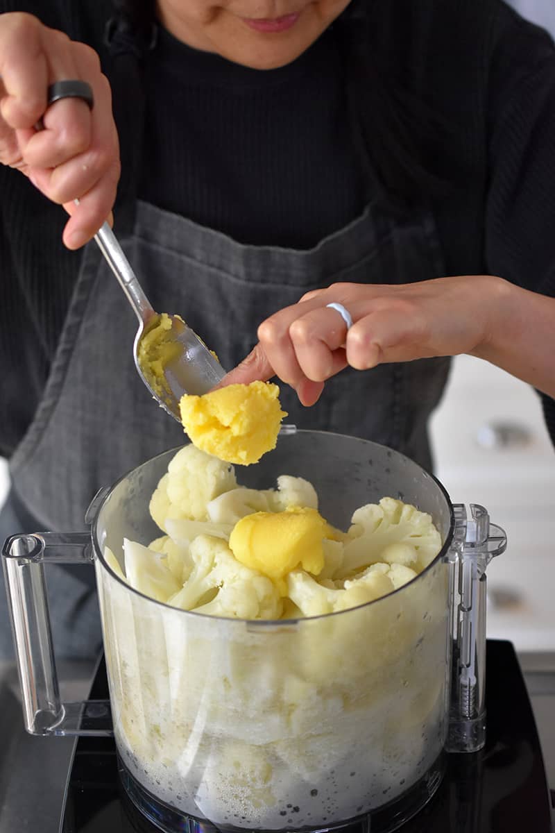 Someone in a gray apron is adding ghee to an open food processor filled with cooked cauliflower florets.