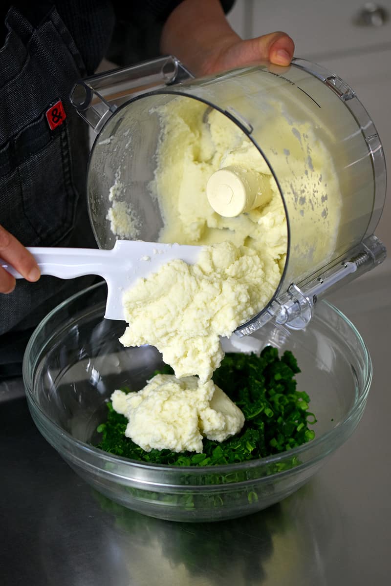 Using a white spatula to spoon cauliflower puree into a large bowl filled with chopped kale and scallions.
