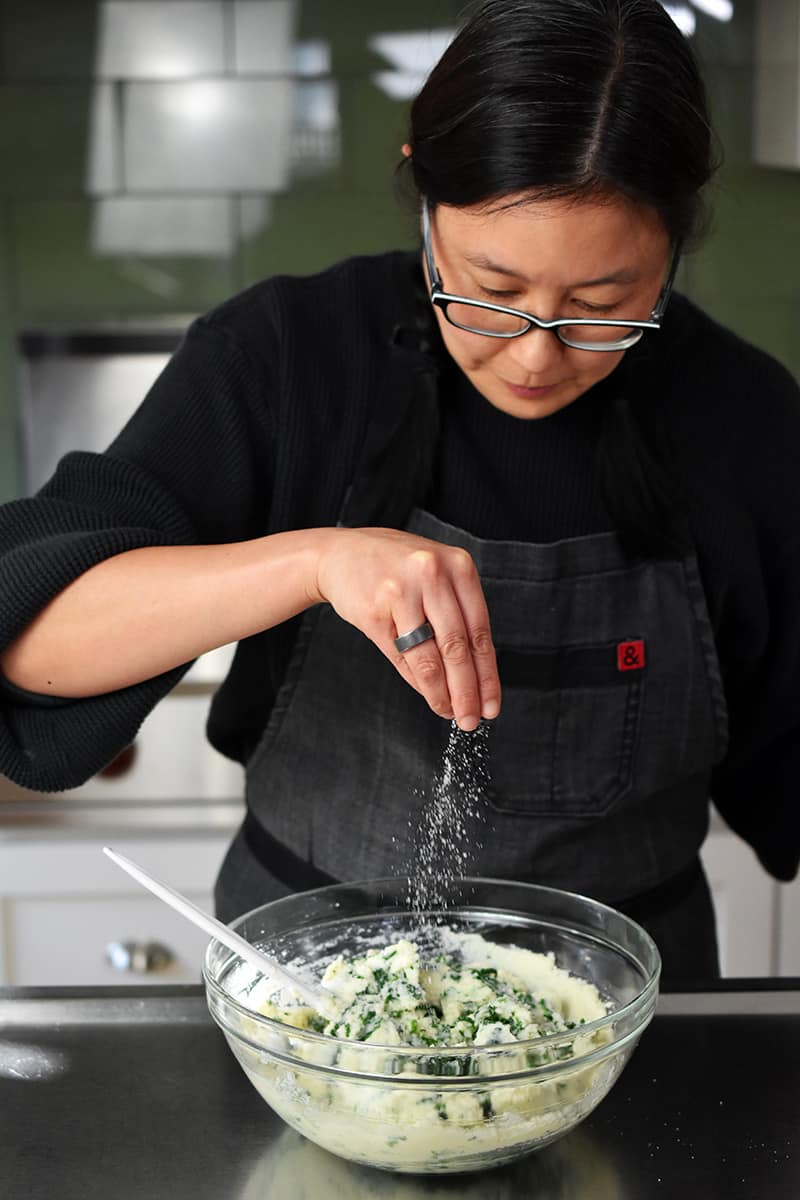 A woman in a gray apron is sprinkling salt on a big bowl filled with cauliflower colcannon