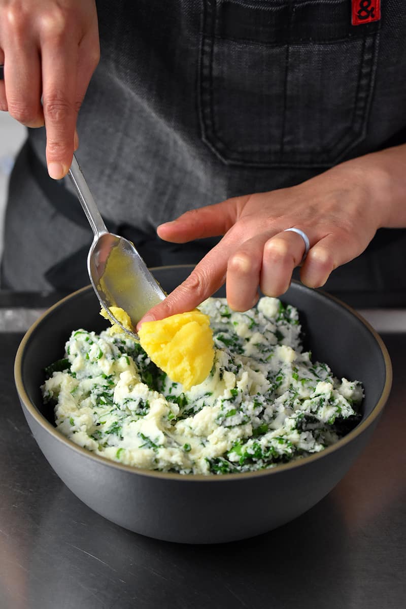 A hand is pushing a dollop of ghee into a bowl of cauliflower colcannon.