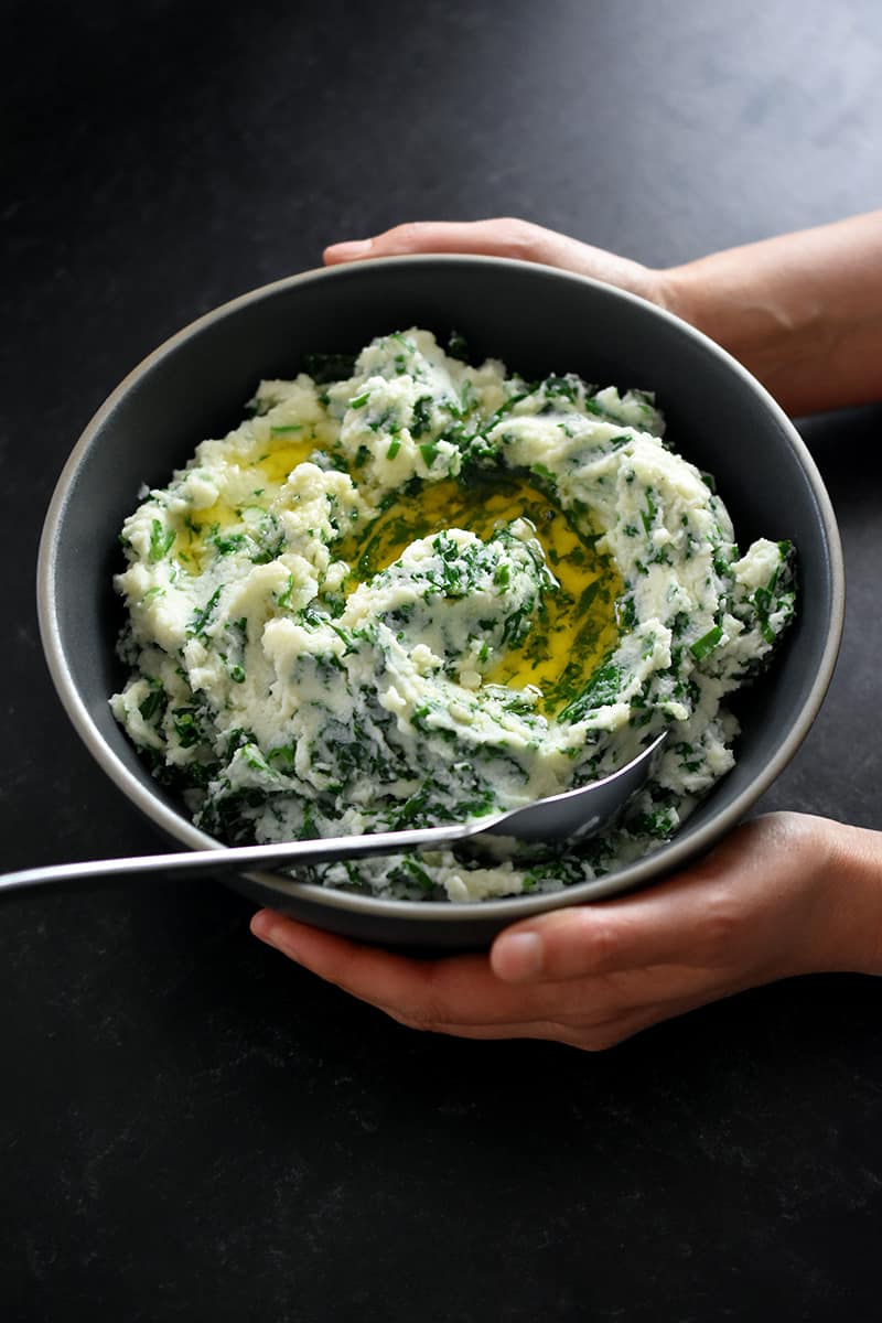 An overhead shot of two hands holding a gray bowl filled with cauliflower colcannon