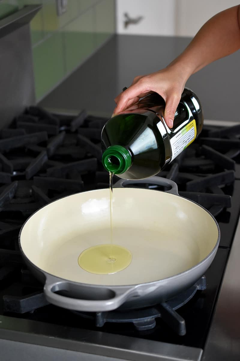 A hand is pouring some avocado oil from a green bottle into a gray ceramic cast iron skillet.