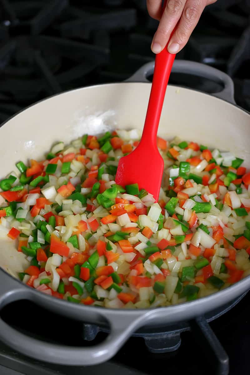 A red silicone spatula is stirring diced onions and peppers in a large Le Creuset braiser.