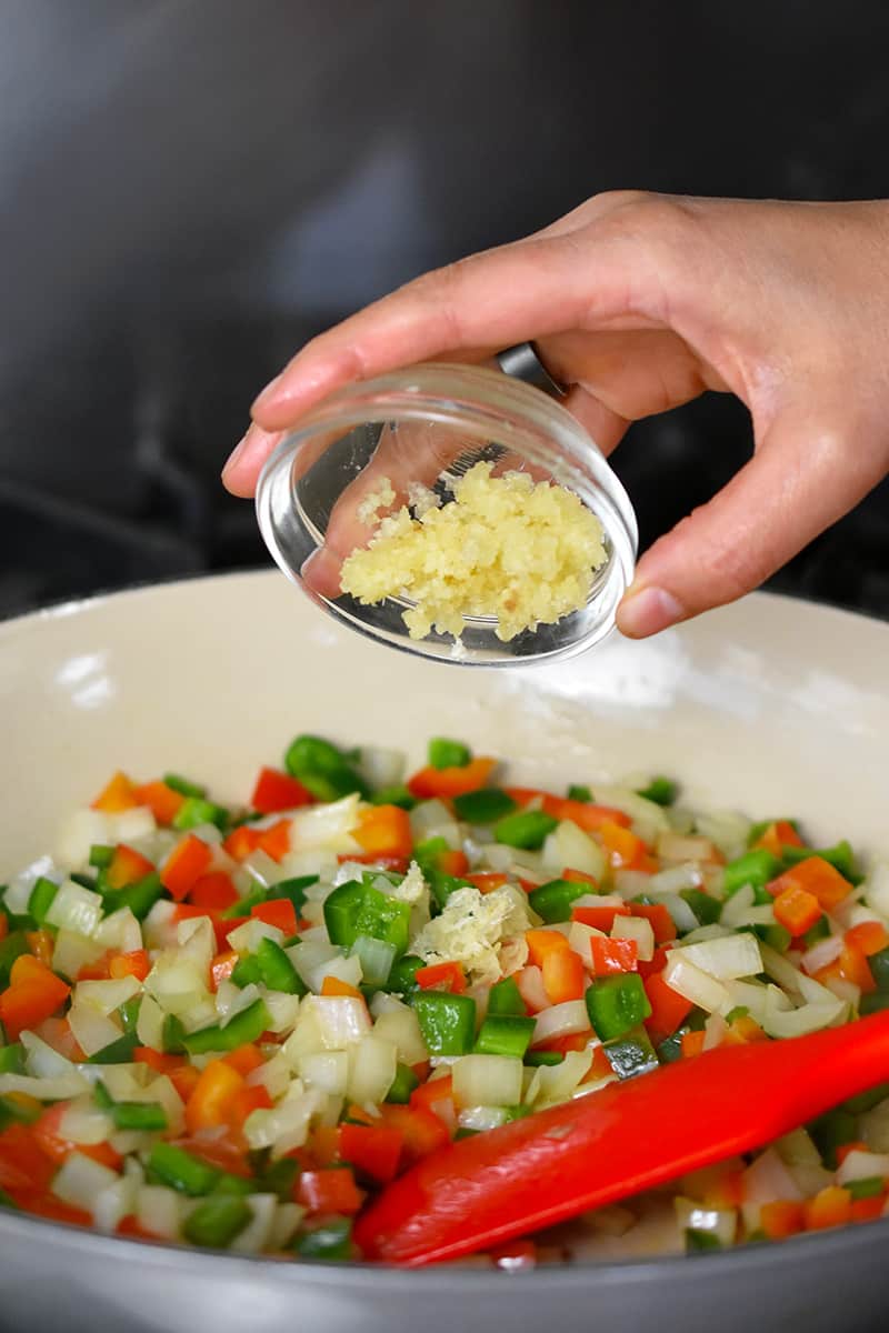 A hand is adding minced garlic from a small glass bowl into the pan of peppers and onions.