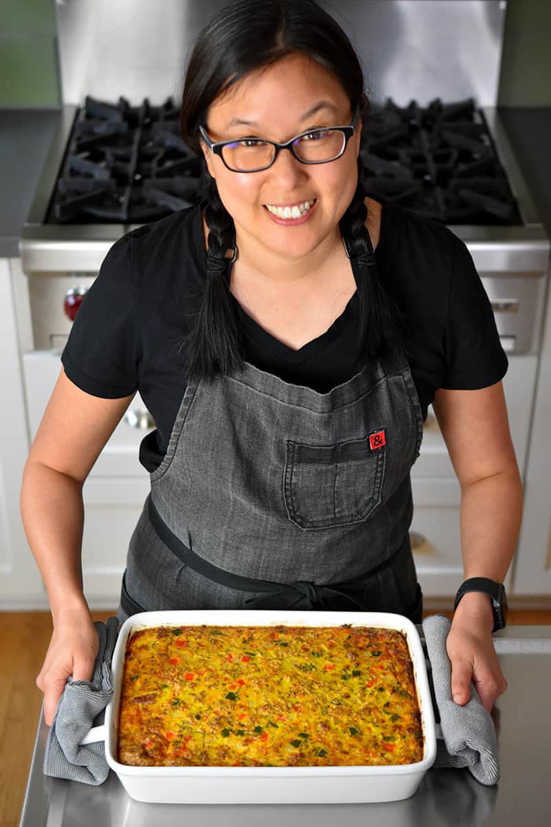 An Asian woman with glasses is smiling and holding the handles of a casserole pan filled with Mexican Breakfast casserole