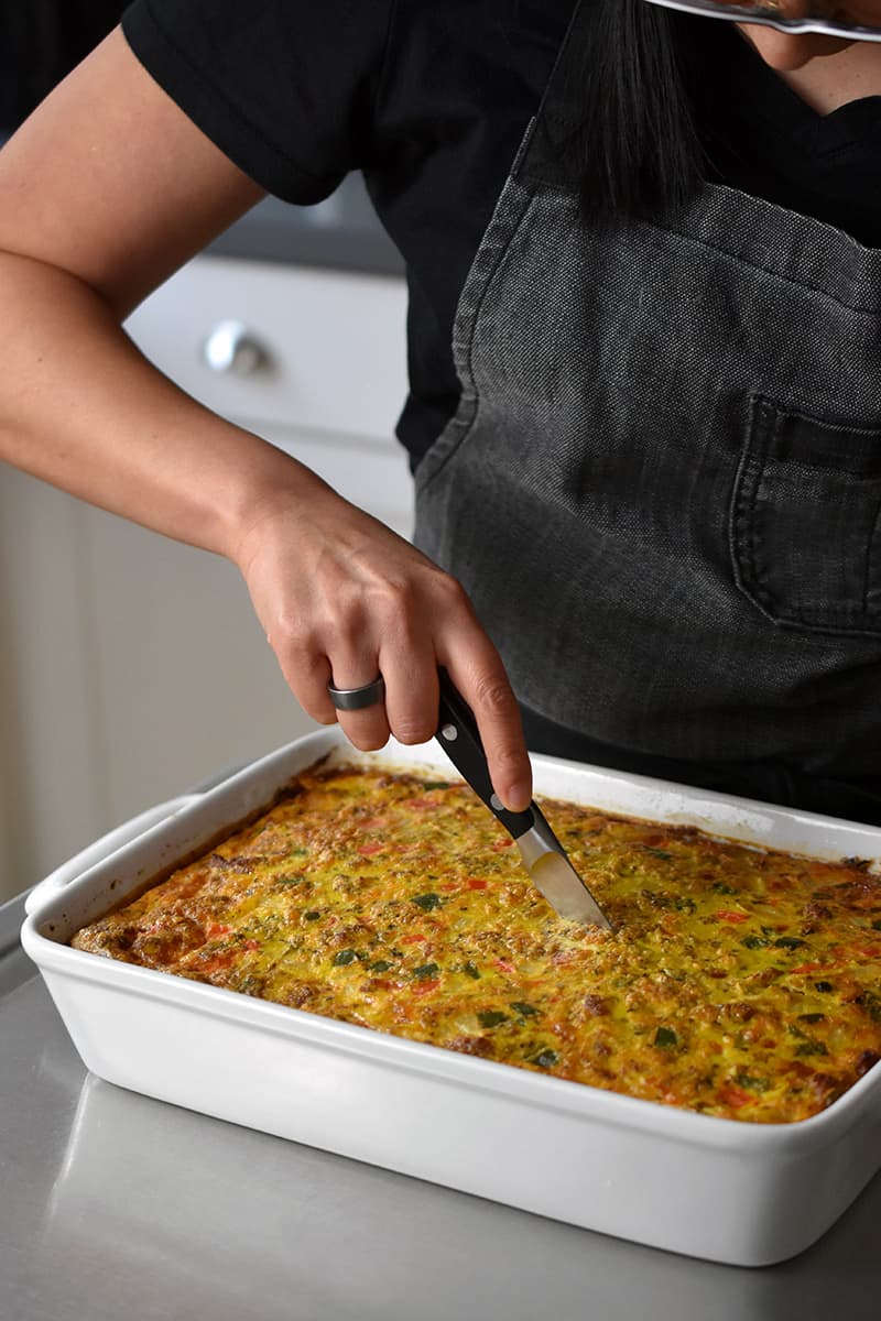 A person in a gray apron is slicing up a Mexican breakfast casserole with a paring knife.
