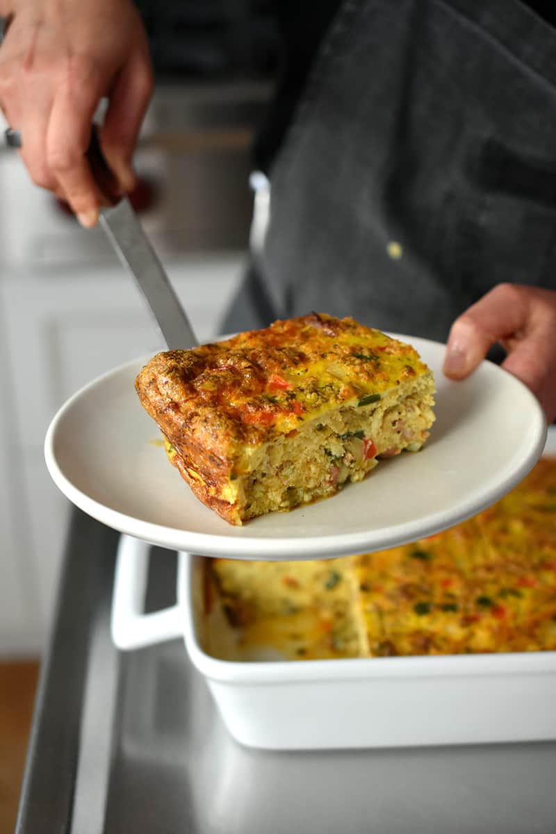 Someone in a gray apron placing a slice of paleo Mexican Breakfast Casserole on a plate.