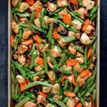 An overhead shot of sheet pan chicken stir-fry right out of the oven. Two hands are holding the edges of the pan with blue and white towels.