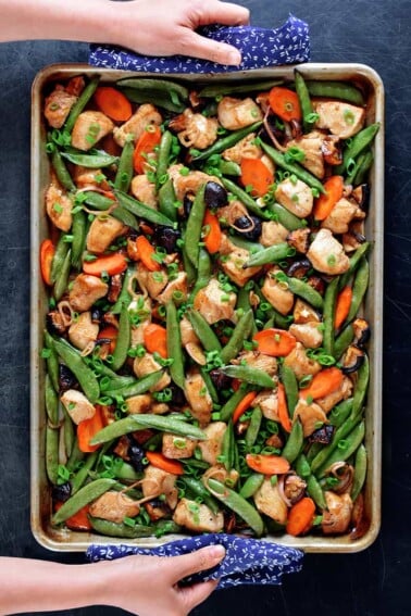An overhead shot of sheet pan chicken stir-fry right out of the oven. Two hands are holding the edges of the pan with blue and white towels.