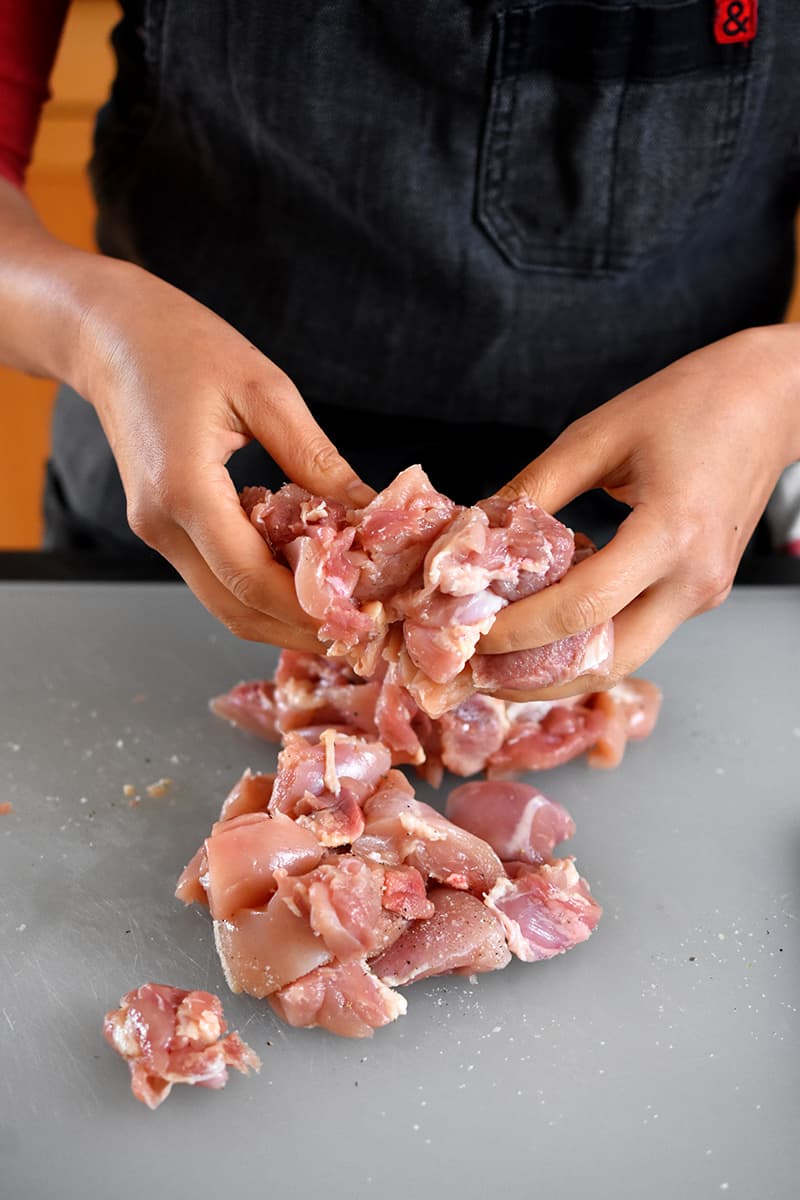Two hands are tossing raw chicken cubes with salt and pepper on a plastic cutting board.