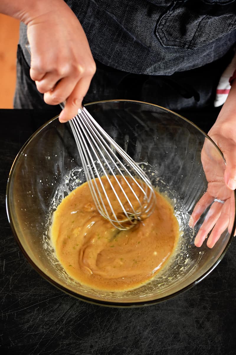 Overhead shot of someone in a gray apron whisking a brown sauce in a clear glass bowl.