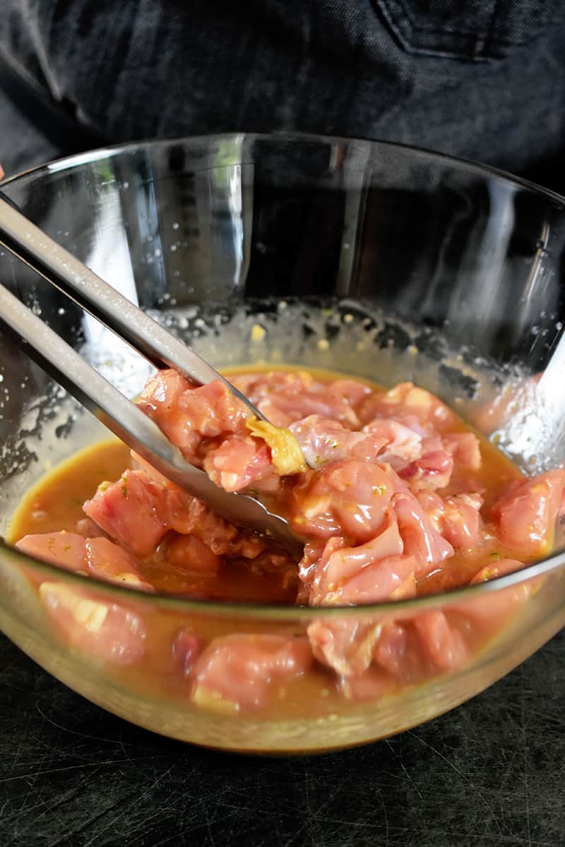 A closeup shot of large glass bowl filled with chicken cubes being tossed in a marinade with a pair of metal tongs