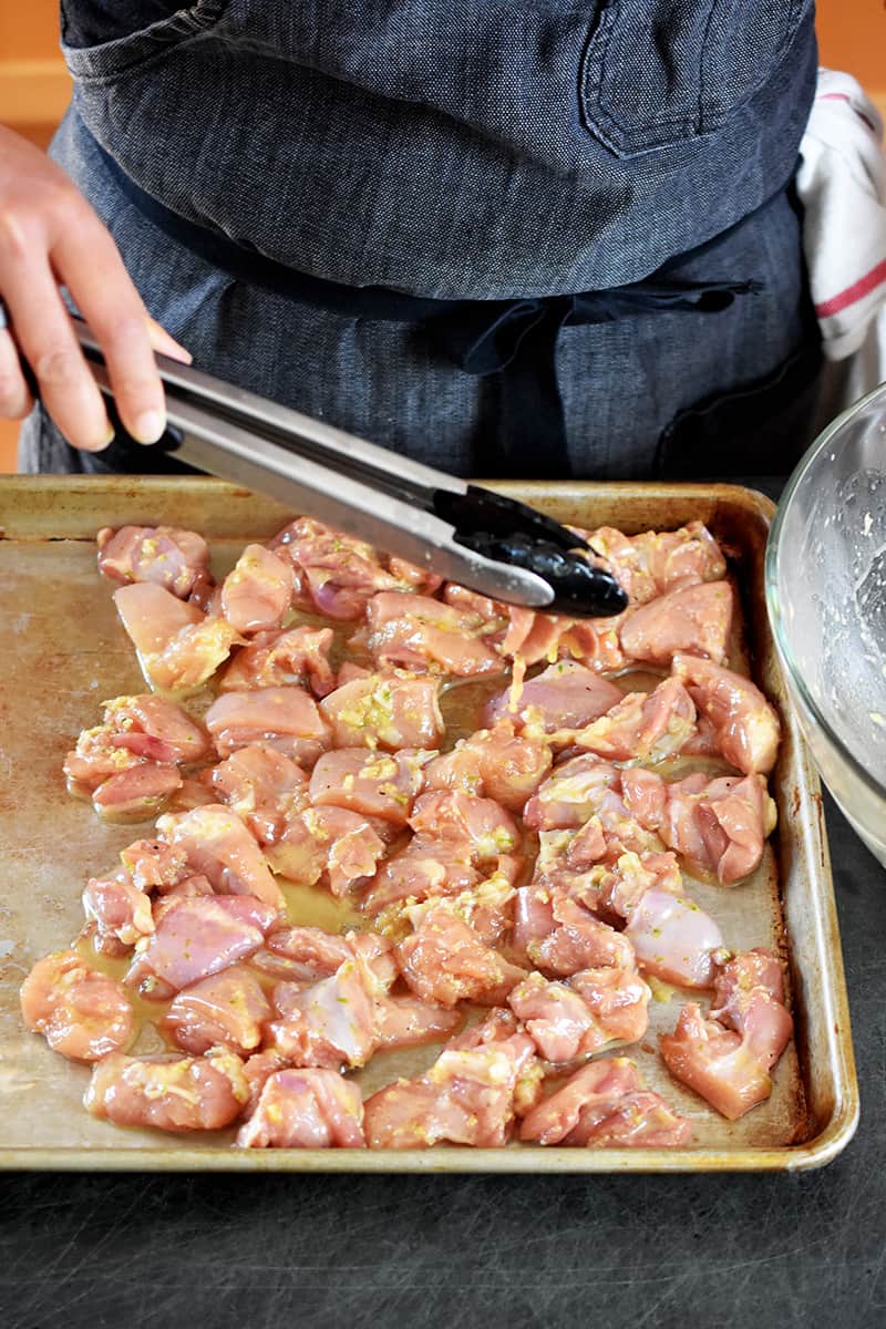 A person is using a pair of tongs to arrange marinated chicken cubes in a single layer on one side of a rimmed sheet pan
