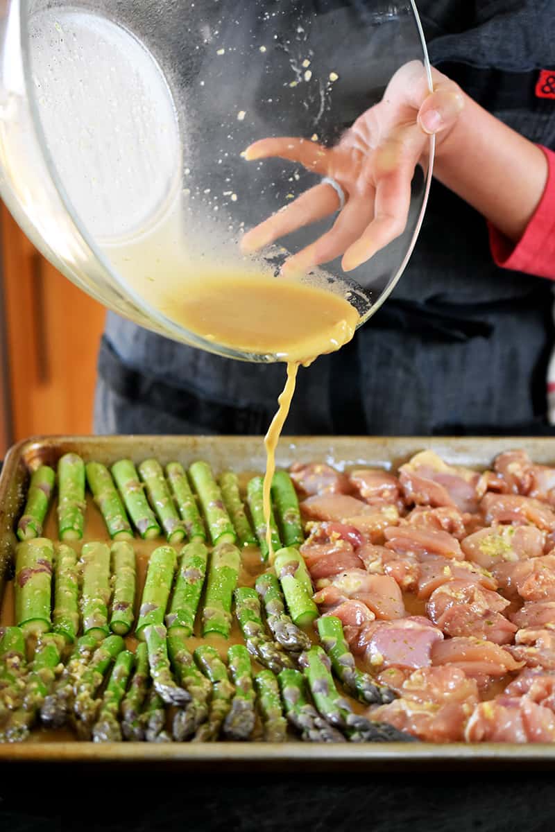 Pouring marinade from a large glass bowl onto a sheet pan filled with cubed chicken and asparagus