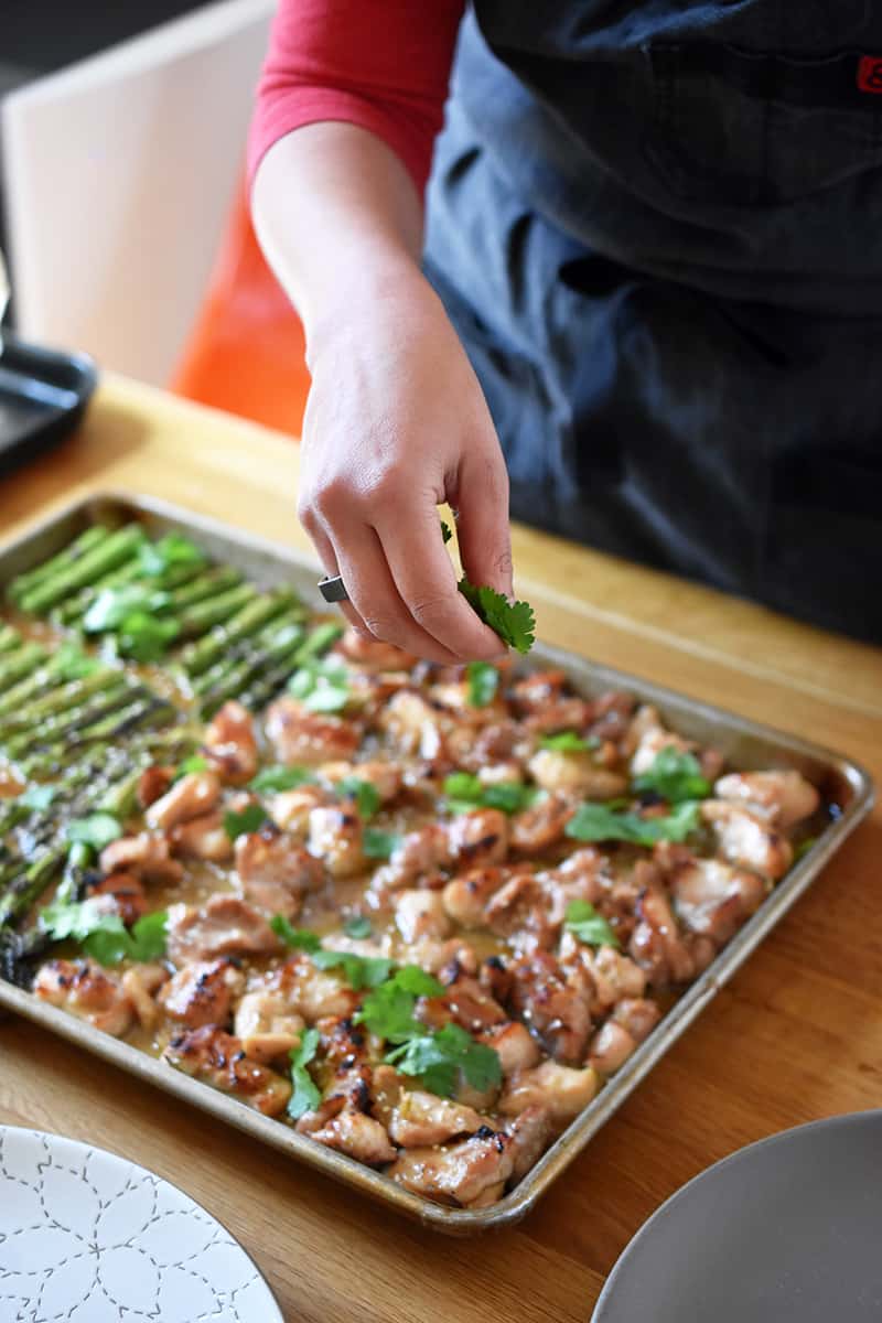 A hand is sprinkling fresh cilantro on a sheet pan filled with chicken and asparagus.