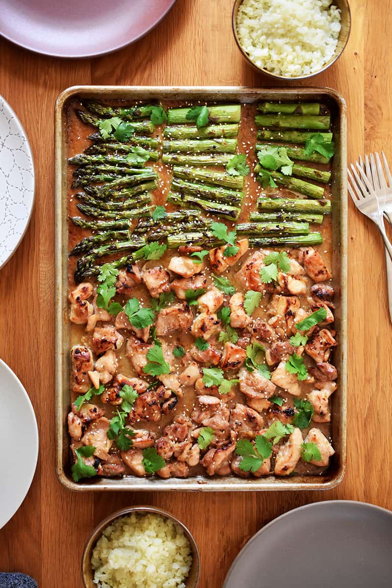An overhead shot of sheet pan chicken and asparagus on a wooden table, next to bowls of cauliflower rice, plates, and forks.