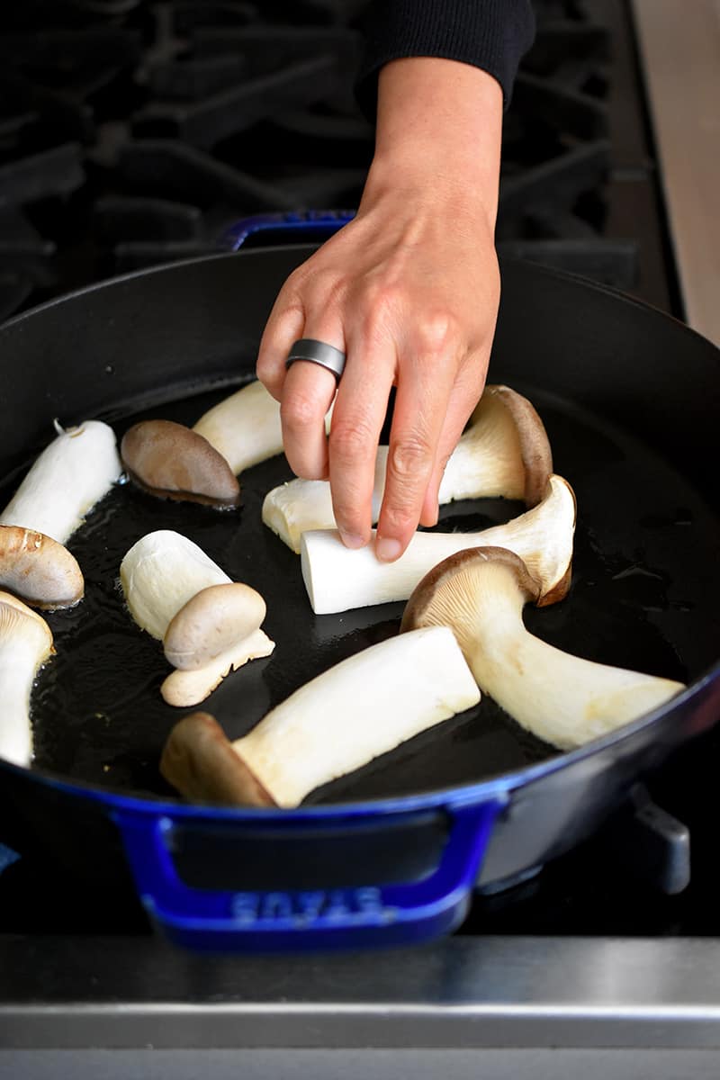 A hand is placing halved king oyster mushrooms cut-side down in a large cast iron skillet