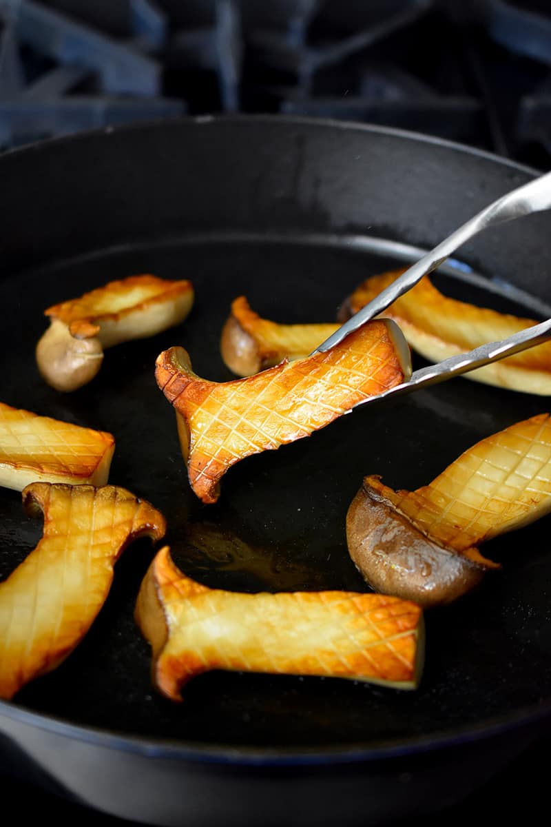 A pair of tongs is flipping golden brown halved king oyster mushrooms in a cast iron pan.
