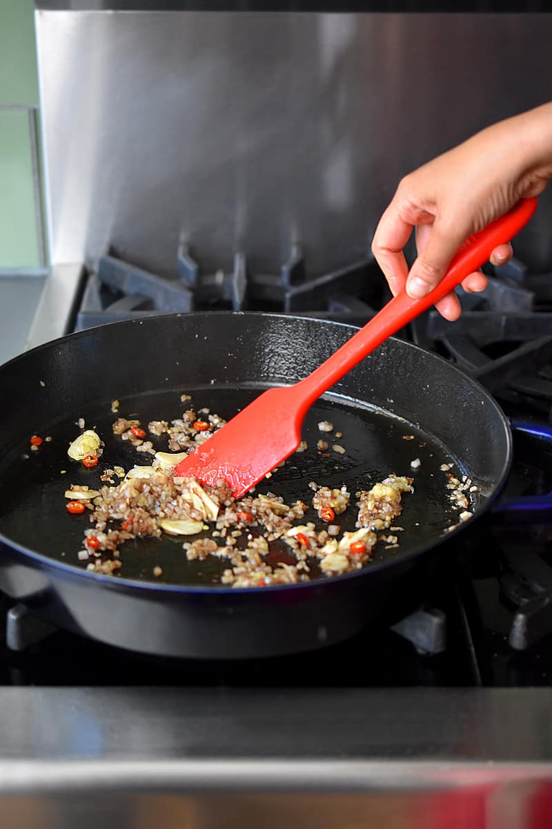 A red spatula is stirring minced shallots, ginger, and garlic in a large cast iron skillet.