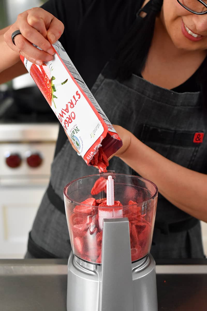 A smiling woman is pouring a package of freeze dried strawberries into a small food processor.