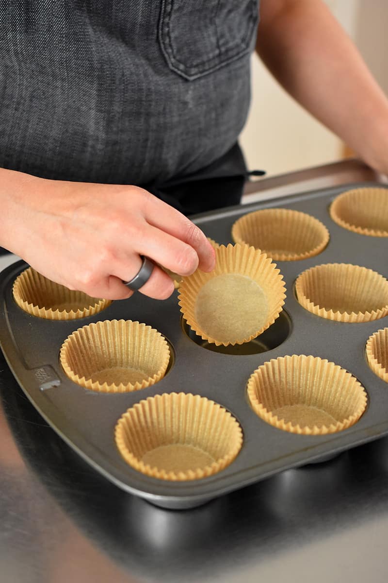 A hand is placing a parchment muffin liner into a muffin tin.