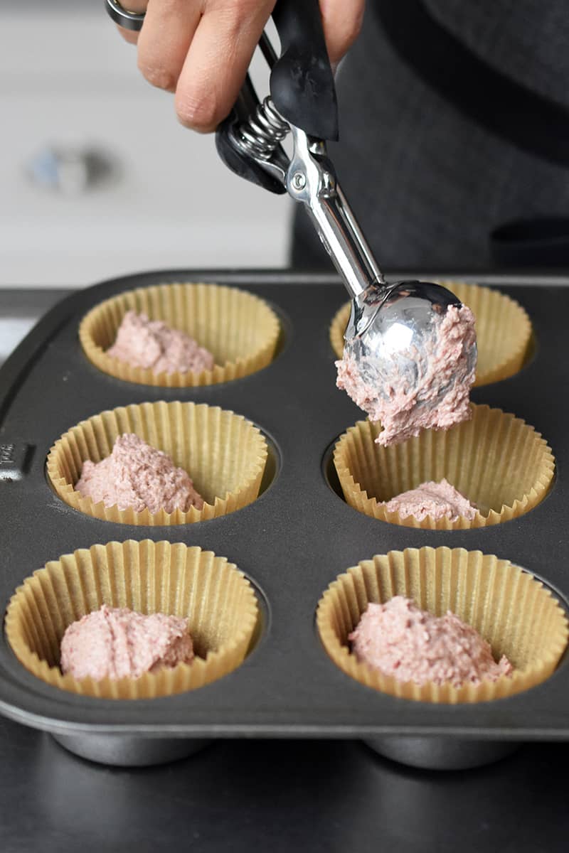 Someone adding a small scoopful of pink batter to the lined muffin pan.