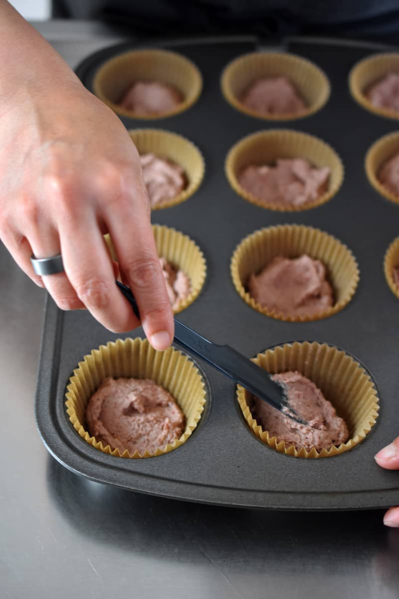 A hand is using a small black spatula to smooth out the pink strawberry muffin batter in the parchment lined muffin pan.