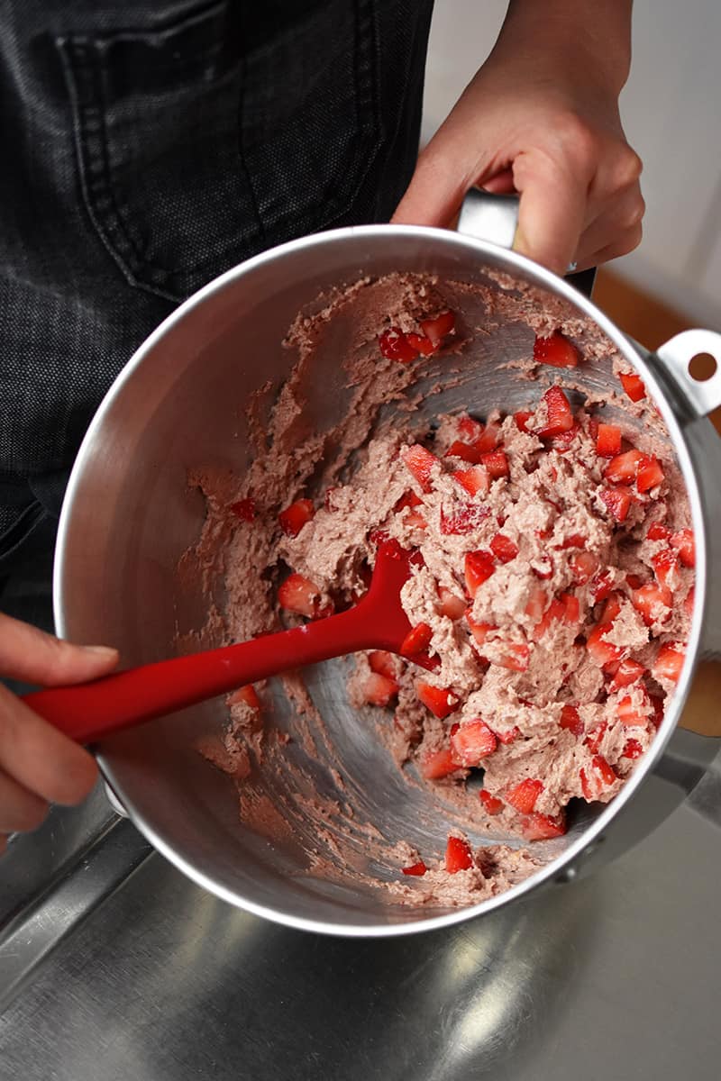 An overhead shot of someone folding in diced fresh strawberries into the pink muffin batter with a red spatula.